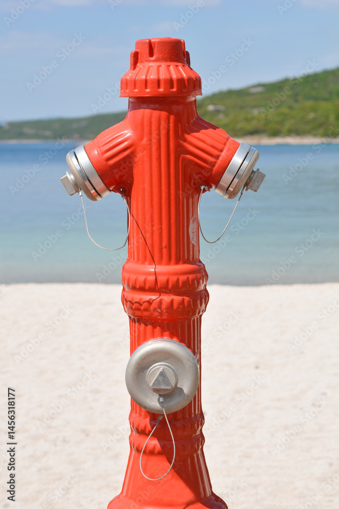 A bright red fire hydrant stands prominently on a sandy surface, with a blurred background of blue sea and a distant green shoreline under a clear sky