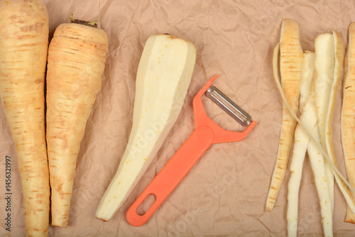 Obraz na płótnie An orange vegetable peeler rests among whole and peeled parsnips on wrinkled beige paper w salonie