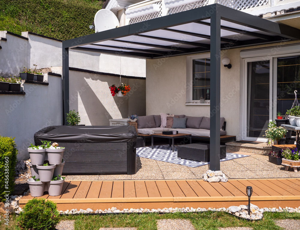 Fototapeta premium Aluminum patio terrace attached to a house with hot tub, sofa, plants, and coffee table on stone tiles, featuring a walkway in front made of composite decking boards.
