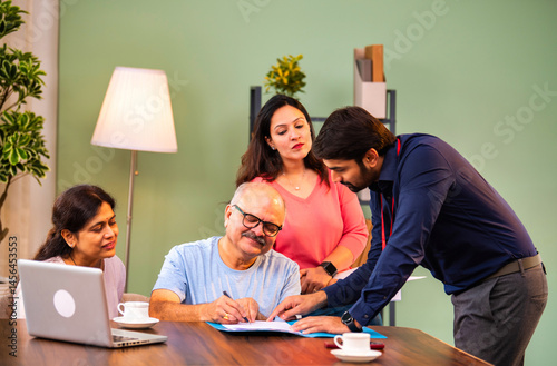 Elderly man signs paperwork with family, guided by young adviser in a modern Indian house