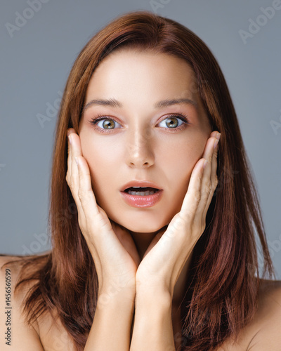 Emotional close-up of a young woman with flawless skin and wide-eyed surprise, hands touching face - perfect for beauty, skincare ads, and expressive lifestyle visuals.