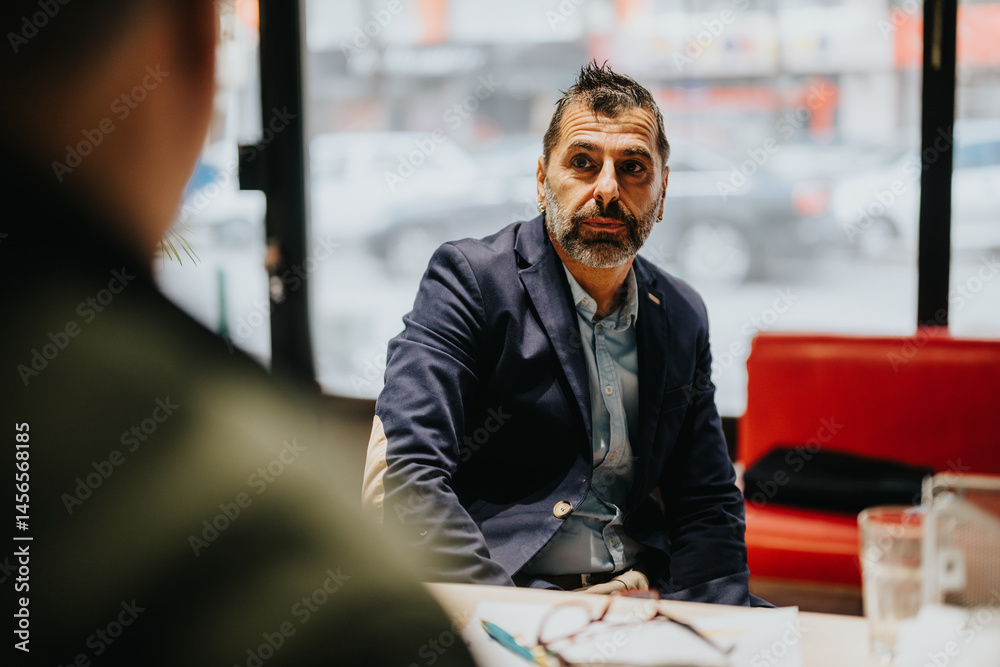 Obraz premium Mature man converses attentively with a colleague inside a modern workplace, surrounded by an urban view, highlighting professionalism and communication during a business meeting.