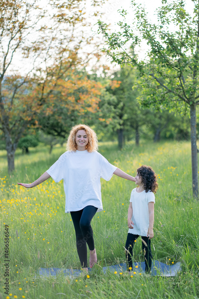 Fototapeta premium Young beautiful mother with her little daughter doing yoga outdoors among the trees at sunset.