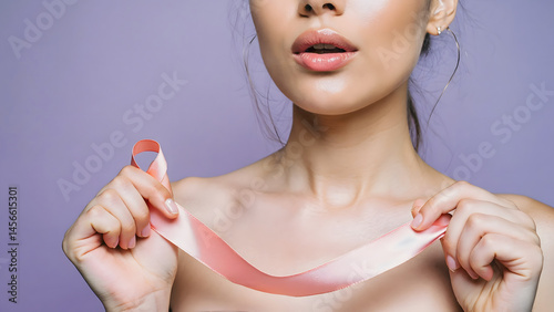 Young woman holding a pink ribbon with one hand looking at camera with pink background. Breast cancer awareness concept.