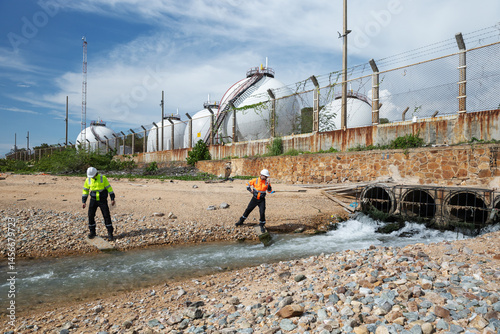 Two engineers assess water discharge from industrial drainage pipes near spherical storage tanks, checking environmental impact and pollution levels in a rocky coastal area.