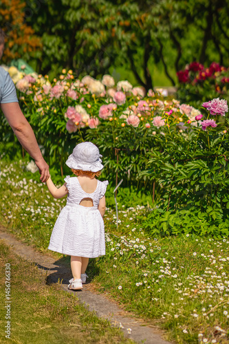 Baby girl walking in flowers park t with father holding her hand. Little girl in white dress walking along blooming garden path holding adult hand. Concept of childhood, safety, tender family moments