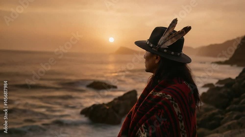 Indigenous person in traditional feathered hat and blanket gazes thoughtfully at the golden ocean sunset from a rocky coastline. Serene, spiritual moment.