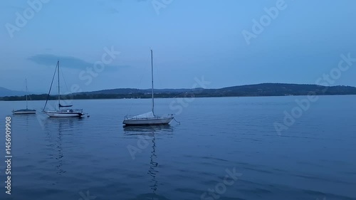 Lake Maggiore, Arona, Italy - May 3 2025: Moored sailboats rock softly near Arona’s harbor at dusk, gentle waves lapping beneath pastel skies above Lake Maggiore’s peaceful expanse.