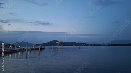 Lake Maggiore, Arona, Italy - May 3 2025: The lake stretches wide beyond old wooden docks near Arona, clouds glowing softly under a fading sky in the tranquil evening hush.