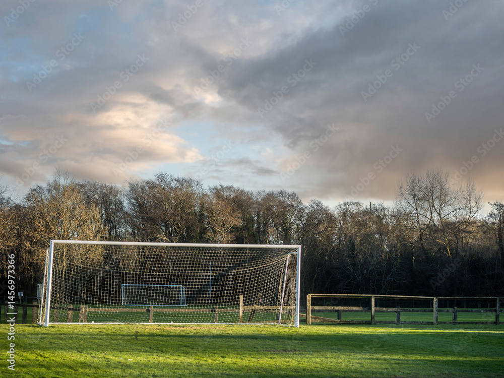 Fototapeta premium A soccer field with a goal and a fence in the background. The sky is cloudy and the grass is green. Football training ground. Popular outdoor sport. Nobody.