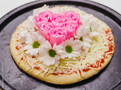 Pink roses in heart love shape and white chamomile flowers on top of a pizza and on dark metal cooking round tray and on white background. Abstract conceptual art. Selective focus.
