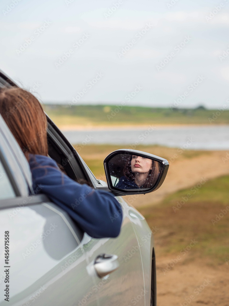 A woman is sitting in a silver car. She is wearing a blue hoodie and gray sweatpants. The scene is quiet and peaceful. Model looking at calm nature scene. Travel to nature