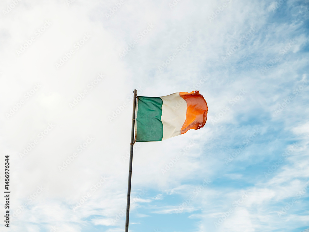 Irish national flag waving on a pole, blue cloudy sky background.