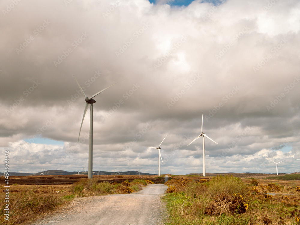 A field of wind turbines with a clear blue cloudy sky in the background. The windmills are tall and spread out across the field. Production of clean green energy. Warm sunny day. Nobody.