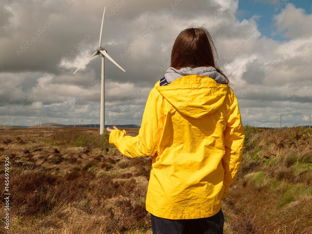 Young teenager girl in yellow jacket, Wind turbines and vast wild fields in the background. Learning clean and green energy production. Warm sunny day.