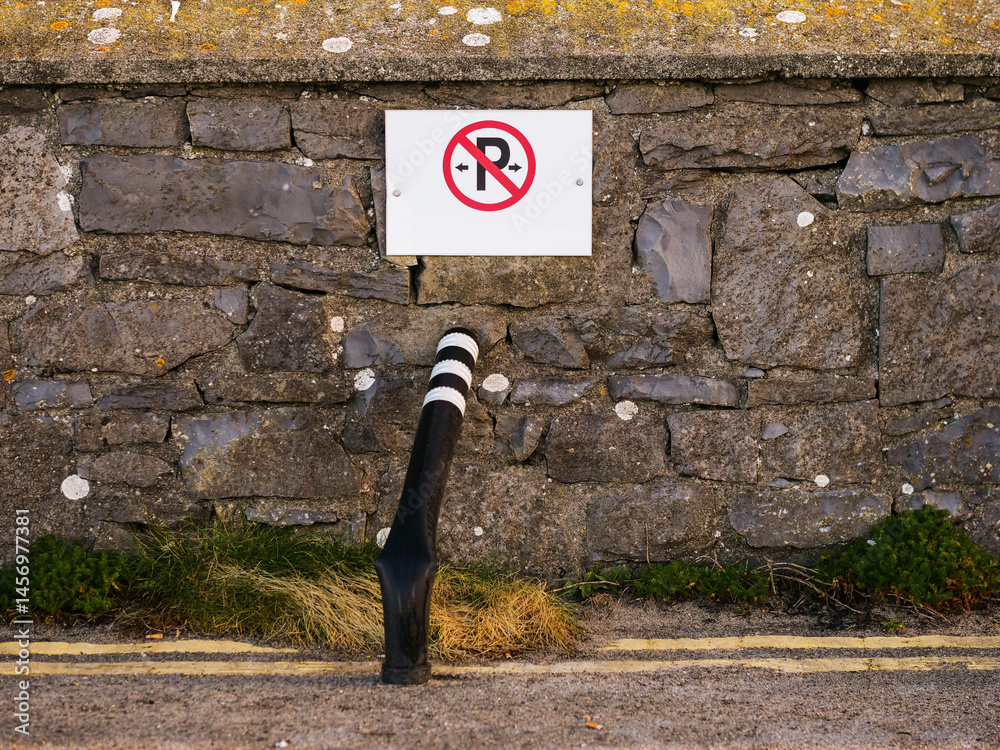 No parking sing on a stone wall and bent and damaged black safety pole with reflective surface by a double yellow line on road surface. Security measures in high risk dangerous area.