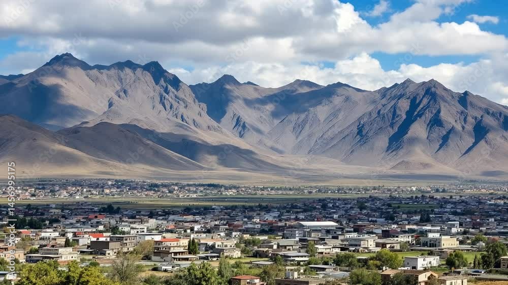 town nestled beneath rugged mountain range under a partly cloudy sky on a sunny day