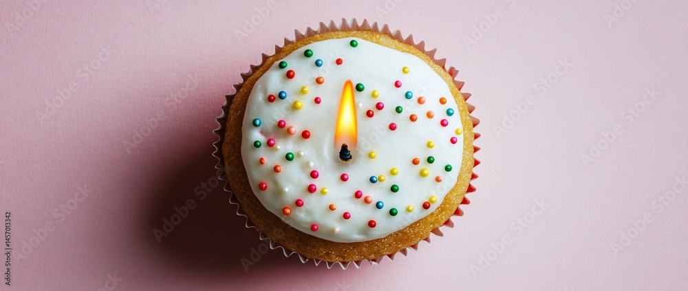 Single cupcake with lit candle and sprinkles on pink background.