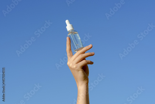 Female hand holding transparent glass mock-up tube of facial serum on blue sky background. Concept of cosmetic products for moisturizing, nourishing and smoothing skin