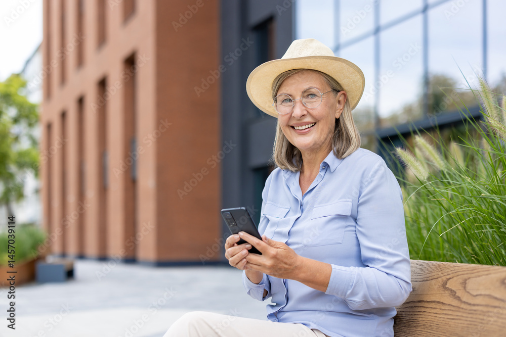 A smiling senior woman using her phone outdoors, wearing a hat and glasses. She is sitting on a bench enjoying the day.