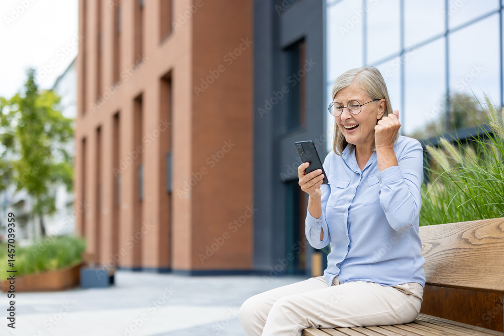 An excited senior woman wearing glasses looks at her phone, celebrating good news while sitting outside. She is happy.