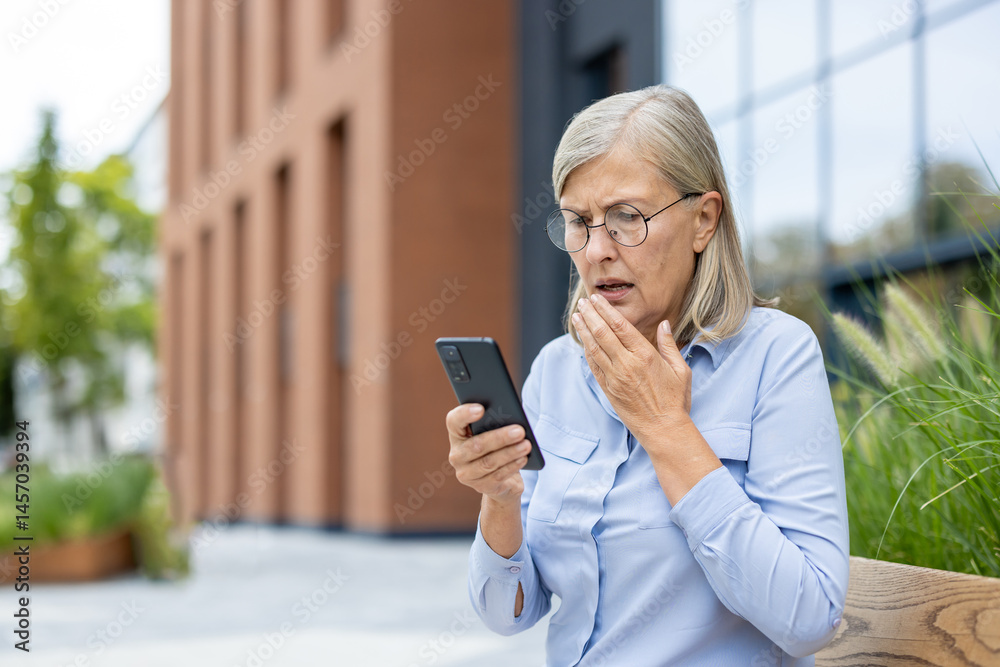 An older woman is looking at her phone with a look of concern on her face, outdoors. She is holding her hand to her mouth as well.