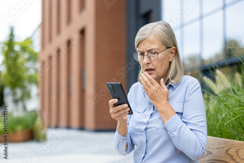An older woman is looking at her phone with a look of concern on her face, outdoors. She is holding her hand to her mouth as well.