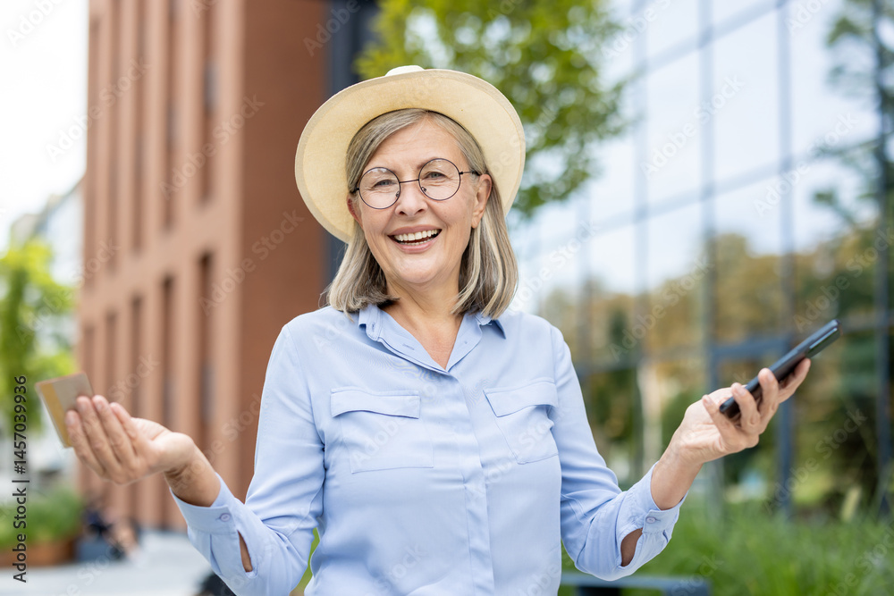 Smiling senior woman holds a credit card and a phone outdoors. She wears a hat, glasses, and a blue shirt near a modern building.