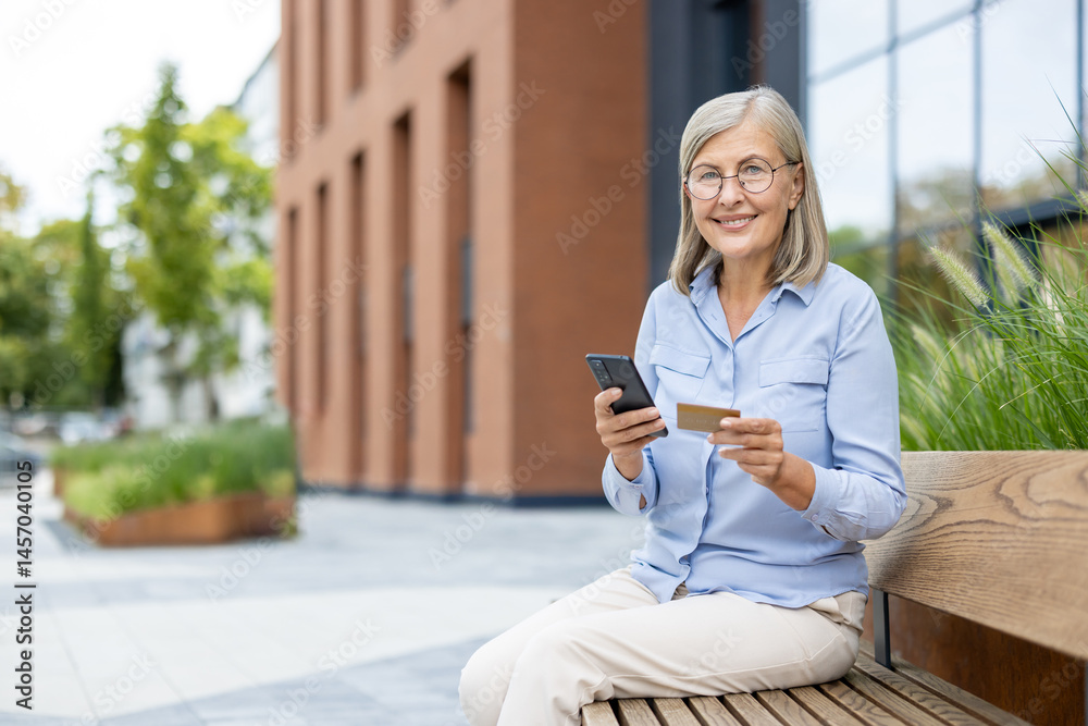 An older woman sits outdoors, smiling while looking at her phone and holding a credit card. She is enjoying a sunny day.