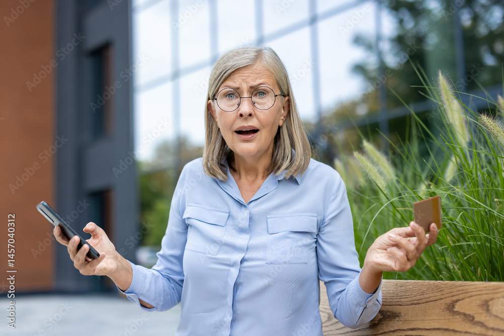 Concerned senior woman holding a credit card and a phone, expressing worry about her online transaction outdoors near an office building.