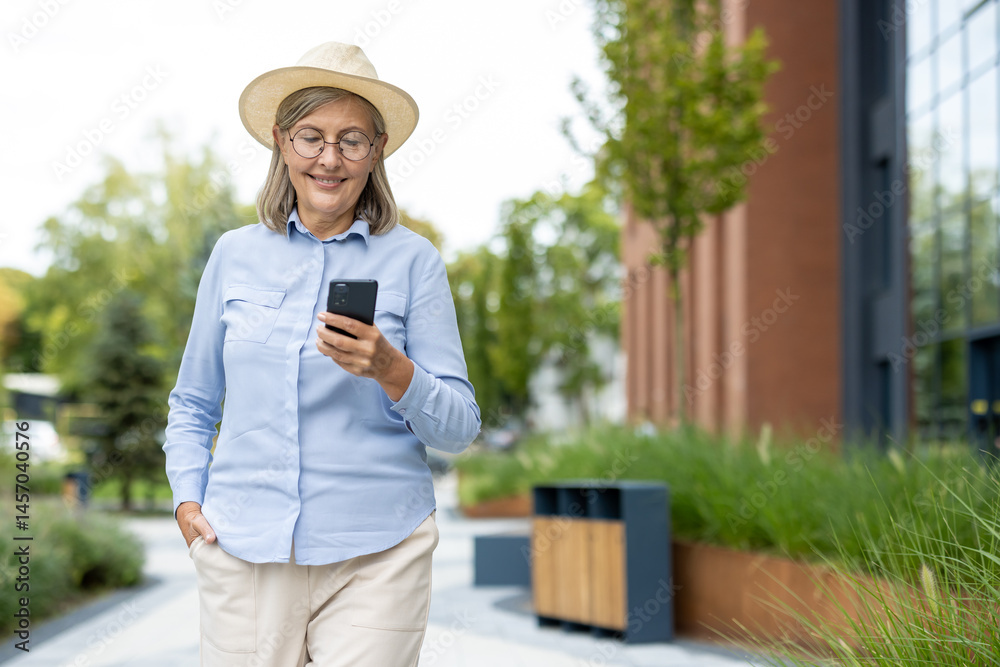 A smiling senior woman in a hat uses her phone while walking outdoors in a modern urban setting, enjoying a sunny day.