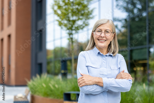 Obraz na płótnie A smiling senior woman with crossed arms poses outdoors near a modern building with glass windows. w salonie