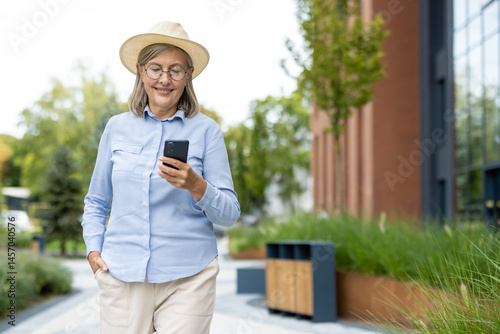 Obraz na płótnie A smiling senior woman in a hat uses her phone while walking outdoors in a modern urban setting, enjoying a sunny day. w salonie