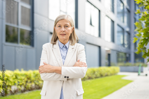 Obraz na płótnie An elegant senior businesswoman stands confidently with arms crossed outdoors near a modern building. She wears a formal outfit. w salonie