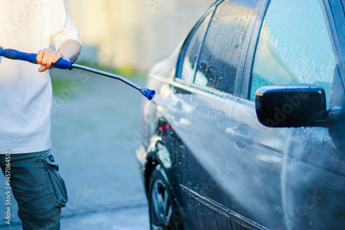 Young man in white T-shirt washing black car with high-pressure washer at self-service car wash, urban background, sunlight reflections on wet surface.

