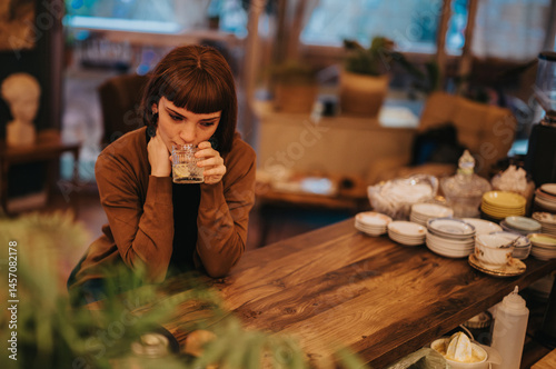 Obraz na płótnie A woman sitting alone at a wooden table, holding a glass, looks thoughtful and introspective. The cozy interior adds a warm atmosphere, contrasting with her contemplative mood. w salonie