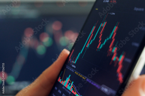 A hand holds a smartphone showing a fluctuating stock chart with red and green candlesticks. Another blurred screen displays similar data in the background.