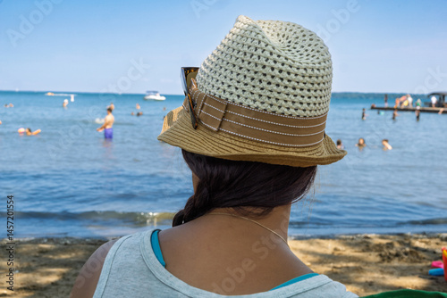 Rear view of a young woman wearing a straw fedora hat on a beach and looking at bathers in the lake