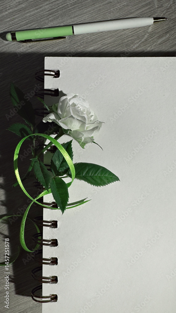 White rose with green ribbon on spiral notebook and pen on wooden surface, perfect for romantic or creative themes