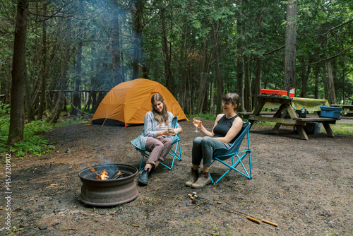 Young LGBTQ couple relaxing by campfire at forest campsite with wine