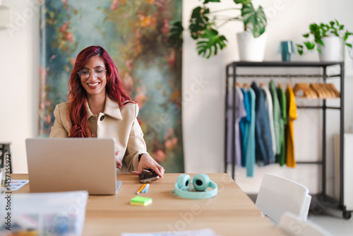 An Arab woman smiles while working on her laptop. She is in a bright, modern office space with plants and a clothing rack.