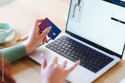 An Arab woman shops online using a laptop and a credit card. She is browsing a fashion website, selecting clothes for purchase. card. She is browsing a Mango website, possibly purchasing a dress.