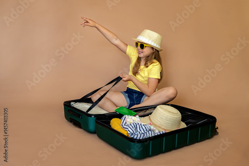 Little girl in yellow glasses and hat sitting in suitcase and pointing direction with cuckoo ready for travel. Background with space for text. Child on vacation or holiday. Travel concept.