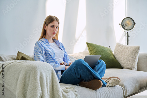 Portrait of young woman sitting on sofa with laptop looking at camera, in home interior