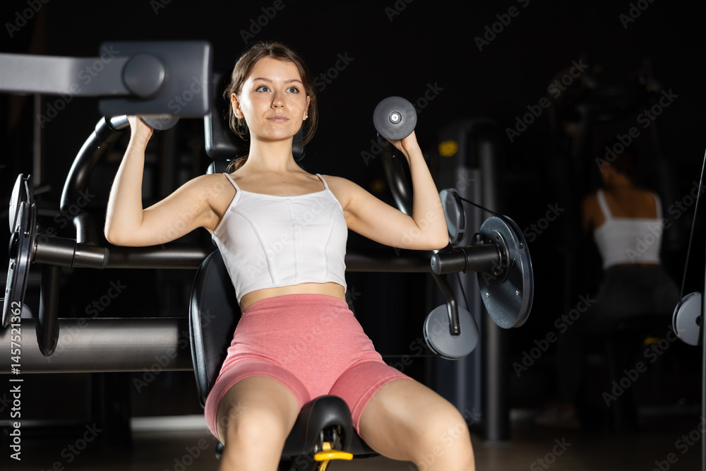 Fototapeta premium Slender athletic girl work out muscles of arms and hands on simulator in fitness club. Gym visitor trains quadriceps during classes at multi-station in wellness center.