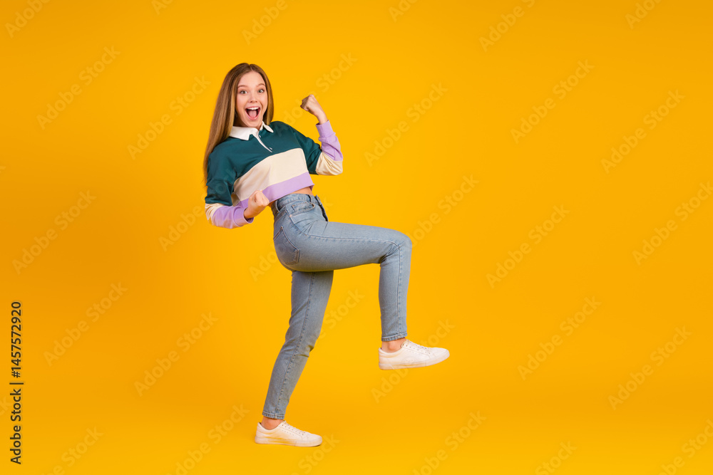 Happy young woman in casual colorful sweatshirt celebrating success against vibrant yellow background