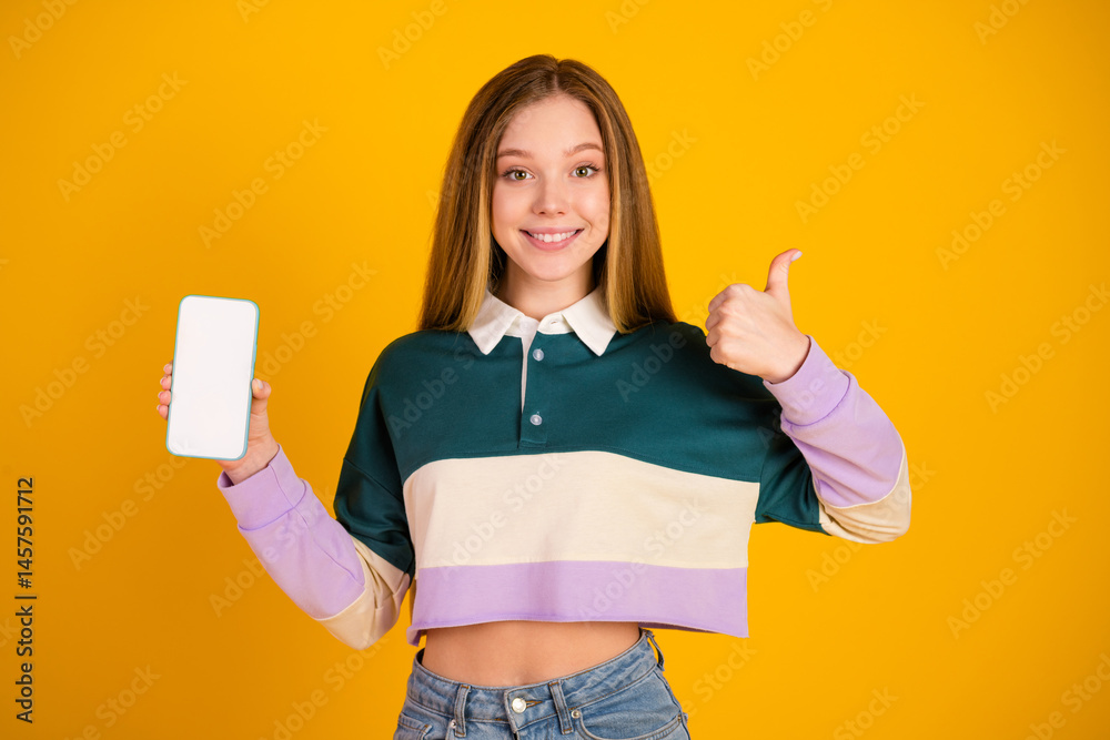 Young woman in stylish casual outfit smiling and showing a smartphone with thumbs up against a yellow background