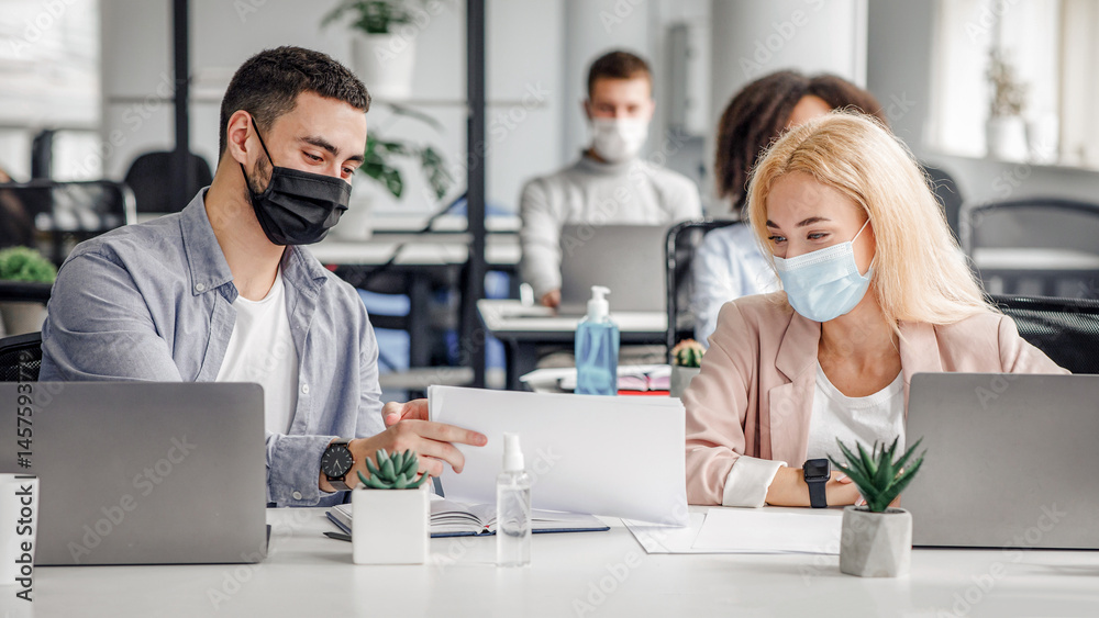 Social distance and work with documents in modern office. Millennial man and woman in protective masks in workplace with laptops and antiseptic, talk after quarantine covid-19, panorama, copy space Social distance and work with documents in modern office. Millennial man and woman in protective masks in workplace with laptops and antiseptic, talk after quarantine covid-19, panorama, copy space