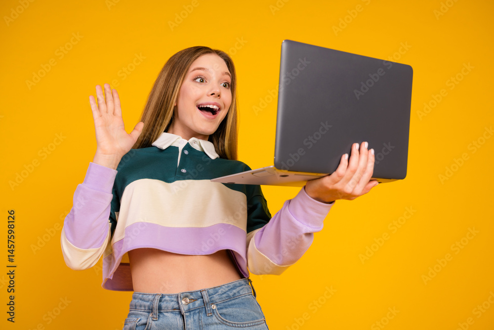 Young woman gesturing happily while holding a laptop against yellow background