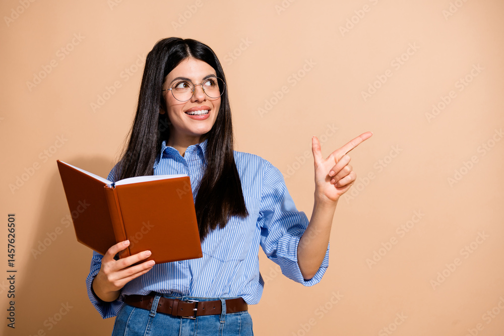Confident young woman in glasses holding a book and pointing to the side with a smile against a beige background Confident young woman in glasses holding a book and pointing to the side with a smile against a beige background